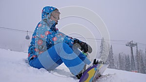 Pretty young woman sitting on mountain peak with her snowboard and enjoying weather during snowfall. Young woman on ski