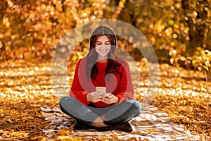 Pretty young woman sitting on picnic blanket at autumn park with headphones and mobile device and listening to music
