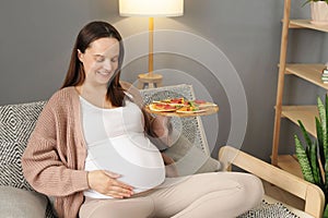 Pretty young woman sitting on sofa holding plate with pizza smiling happily touching her belly enjoying unhealthy snack posing in