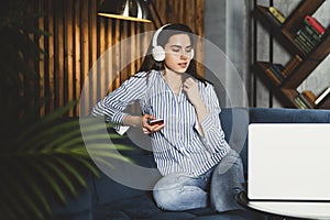 Pretty young woman sitting on sofa and listening to music on smartphone. Girl in earphones streaming songs online in social