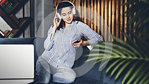 Pretty young woman sitting on sofa and listening to music on smartphone. Girl in earphones streaming songs online in social