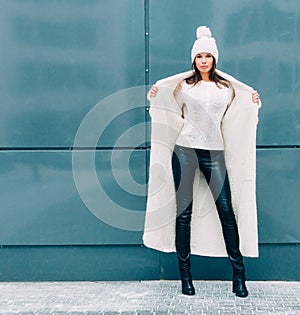 Pretty young woman smiling in a knitted hat and a coat on a grey wall background of the Europe city. Streetstyle.