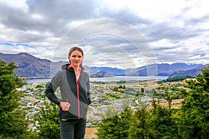 Pretty young woman is staying on the mountain with Panoramic view of lake Wanaka town at the back New Zealand