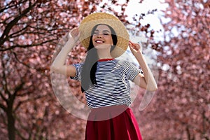 Pretty young woman with straw hat near beautiful blossoming trees outdoors. Stylish spring look