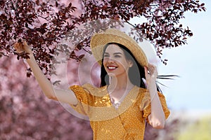 Pretty young woman with straw hat near beautiful blossoming trees outdoors. Stylish spring look