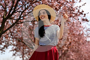 Pretty young woman with straw hat near blossoming trees outdoors. Stylish spring look