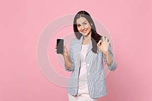 Pretty young woman in striped jacket showing OK gesture holding mobile phone with blank empty screen isolated on pink
