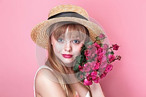 Pretty young woman in summer dress and straw hat holding bouquet of roses on pink background