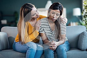 Pretty young woman supporting and comforting her sad friend while sitting on the sofa at home