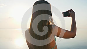 Pretty Young Woman Taking Pictures with her Smartphone at Sunset on the Beach near the Sea on Vacation. Slow Motion