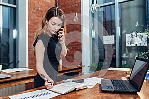 Pretty young woman talking on phone counting using a calculator working at office standing at desk