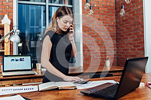 Pretty young woman talking on phone counting using a calculator working at office standing at desk