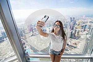 Pretty young woman tourist takes selfie portrait at the top of Burj Khalifa. Beautiful female student takes photo for travel blog