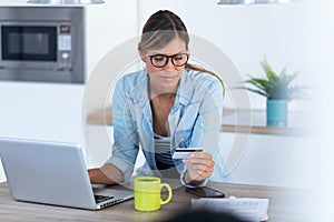 Pretty young woman using her laptpo for shopping online and paying with credit card while sitting in the kitchen at home