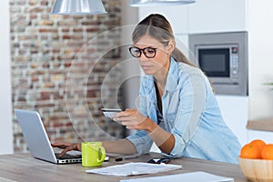 Pretty young woman using her laptpo for shopping online and paying with credit card while sitting in the kitchen at home