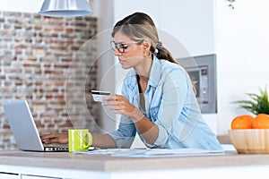 Pretty young woman using her laptpo for shopping online and paying with credit card while sitting in the kitchen at home