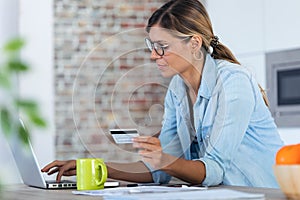 Pretty young woman using her laptpo for shopping online and paying with credit card while sitting in the kitchen at home