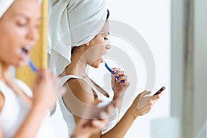 Pretty young woman using her mobile phone while brushing her teeth in the bathroom at home