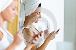 Pretty young woman using her mobile phone while brushing her teeth in the bathroom at home