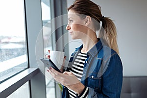 Pretty young woman using her mobile phone while drinking coffee near to the window in the living room at home