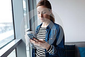 Pretty young woman using her mobile phone while drinking coffee near to the window in the living room at home