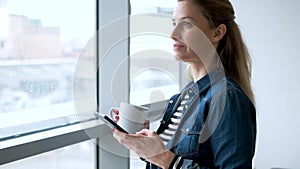 Pretty young woman using her mobile phone while drinking coffee near to the window in the living room at home.