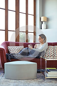 Pretty young woman using her mobile phone while drinking coffee on sofa at home