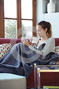 Pretty young woman using her mobile phone while drinking coffee on sofa at home