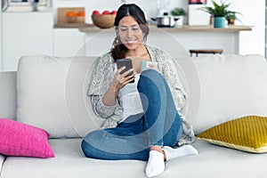 Pretty young woman using her mobile phone while drinking coffee on sofa at home