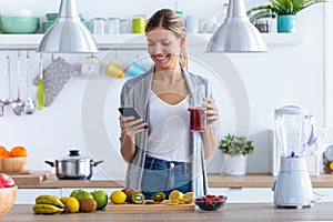 Pretty young woman using her mobile phone while drinking fruit juice in the kitchen at home