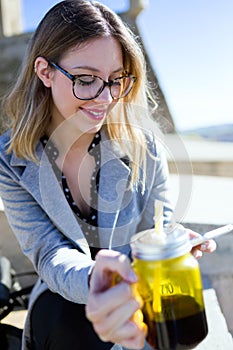 Pretty young woman using her mobile phone while drinking juice in the street.