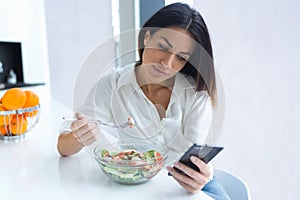 Pretty young woman using her mobile phone while eating salad in the kitchen at home