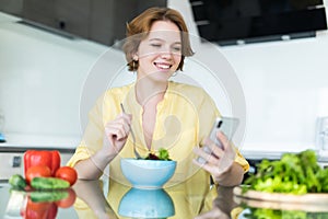 Pretty young woman using her mobile phone while eating salad in the kitchen at home