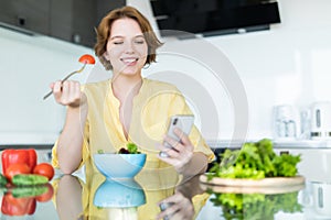 Pretty young woman using her mobile phone while eating salad in the kitchen at home