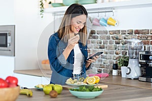 Pretty young woman using her mobile phone while eating a slice of banana in the kitchen at home