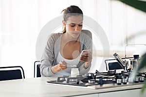 Pretty young woman using her mobile phone while eating yogurt in the kitchen at home