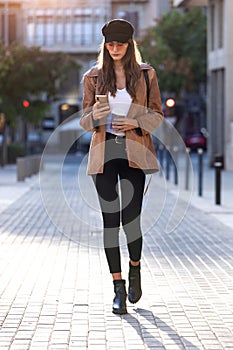 Pretty young woman using her mobile phone while holding a coffee and standing in the street