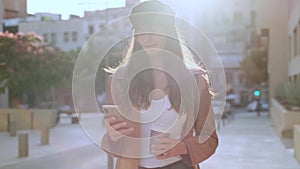 Pretty young woman using her mobile phone while holding a coffee and standing in the street.