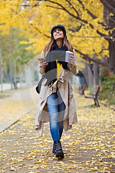 Pretty young woman using her mobile phone while looking to sky and walking through the park during autumn