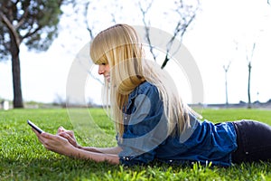 Pretty young woman using her mobile phone while lying on grass in the park.