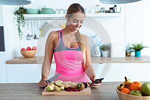 Pretty young woman using her mobile phone while preparing detox juice in the kitchen at home