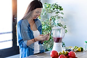 Pretty young woman using her mobile phone while preparing fruit smoothie in the kitchen at home