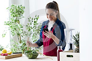 Pretty young woman using her mobile phone while preparing salad in the kitchen at home.