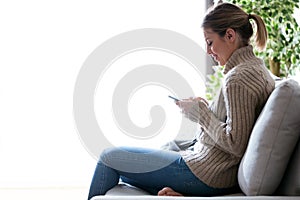 Pretty young woman using her mobile phone while sitting on sofa at home.