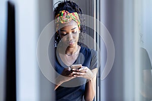 Pretty young woman using her mobile phone while standing next to the window at home