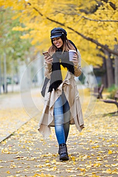 Pretty young woman using her mobile phone while walking through the park during autumn