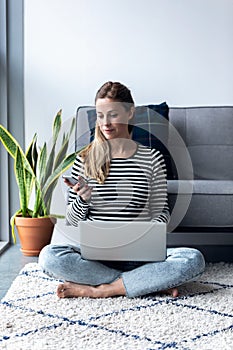 Pretty young woman using her mobile phone while working with laptop sitting on the floor at home