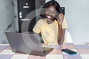 Pretty young african woman using laptop and holding credit card in kitchen