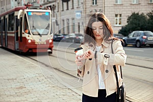 Pretty young woman waiting for train or tram as arrival, checking the time on smartwatch, drinking takeout coffee cup.
