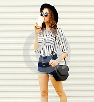 Pretty young woman walking with coffee cup in black round hat, shorts, white striped shirt on white wall